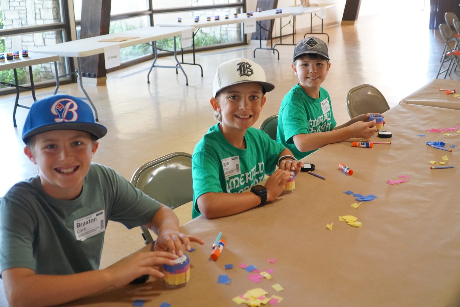 Three smiling boys in Vacation Bible School shirts working on a craft at a table